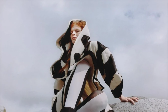 Model with red hair wearing a black and white hooded patterned coat against a cloudy sky
