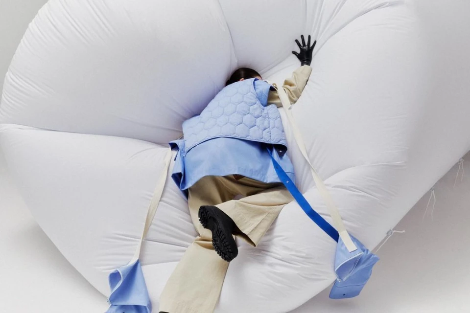 Adult female in blue jacket on puffy white object, wearing black gloves, tan pants and black shoes in a light studio setting.