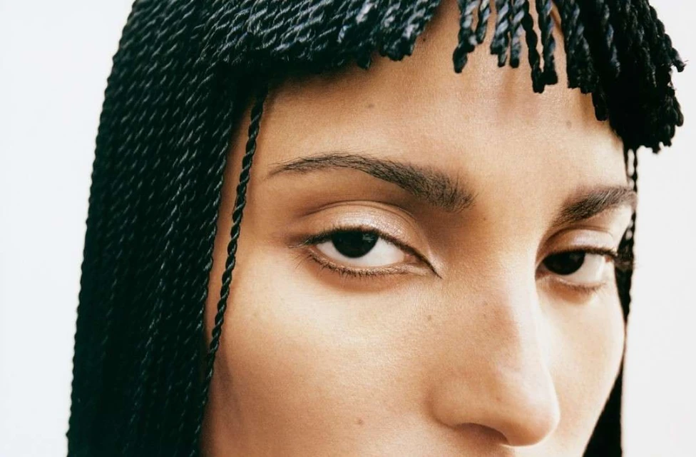 Close-up portrait of a woman's face with black braided hair against a white background, emphasizing her eyes and skin texture.