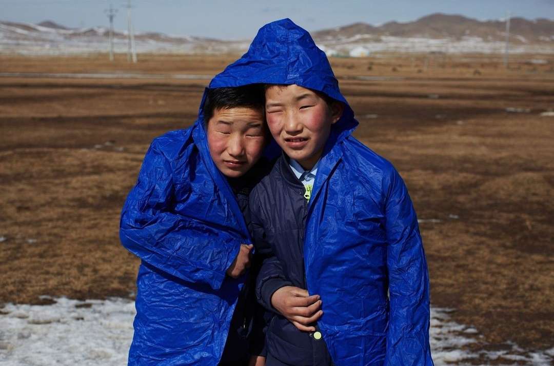 Two young boys in blue hooded raincoats huddle together outside in a field with mountains in the distance. Landscape orientation.