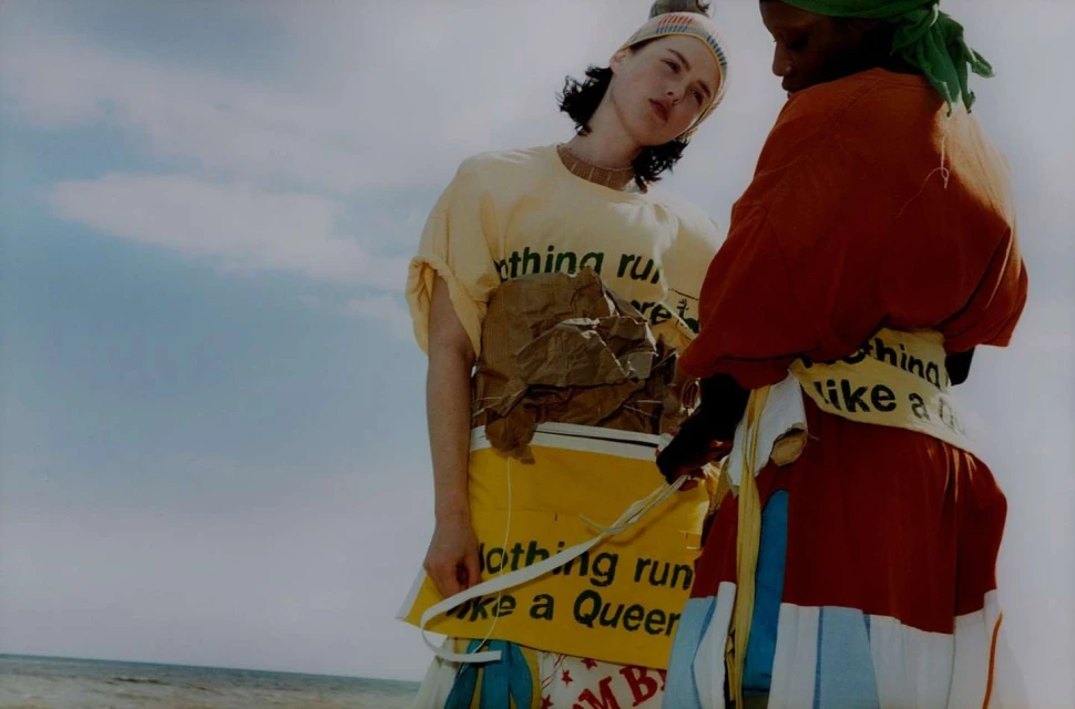 Two models in unique fashion stand on a flat surface under a blue sky, one in yellow and the other in red, styled with text.