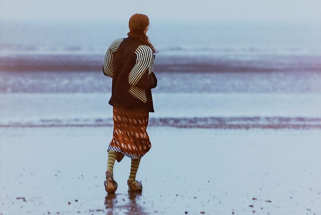 Rear view of a person with long reddish hair walking on a wet sandy beach wearing a patterned skirt and striped sleeves.