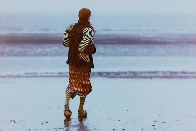 Rear view of a person with long reddish hair walking on a wet sandy beach wearing a patterned skirt and striped sleeves.