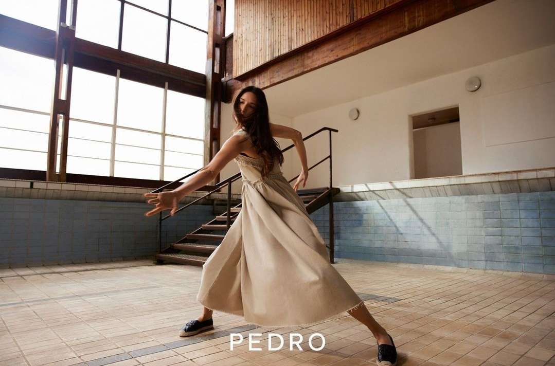 A slender woman in a dress strikes a dance pose indoors with an open geometric window and brown tile flooring, in a medium shot.