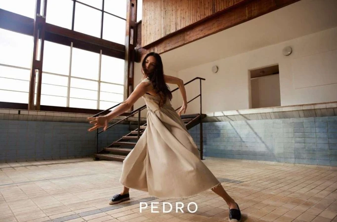 A slender woman in a dress strikes a dance pose indoors with an open geometric window and brown tile flooring, in a medium shot.