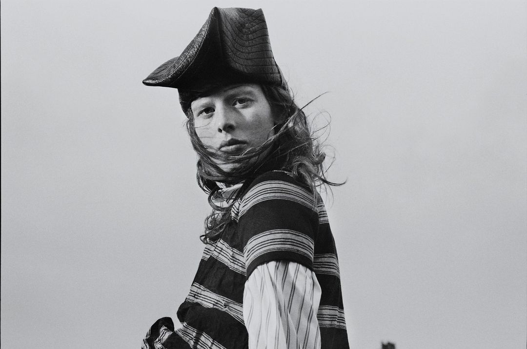 Black and white portrait of a person with long hair wearing a striped shirt and a tricorn hat against a plain, overcast sky.