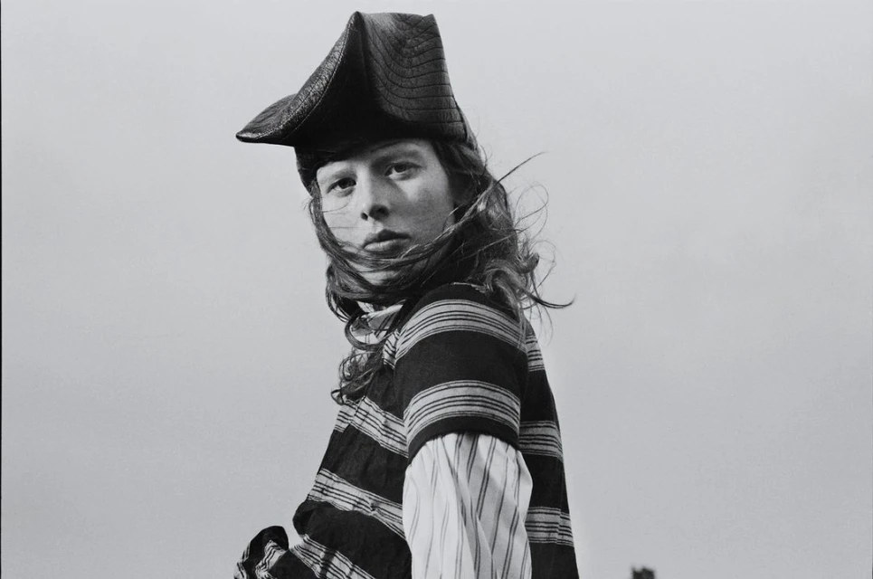 Black and white portrait of a person with long hair wearing a striped shirt and a tricorn hat against a plain, overcast sky.