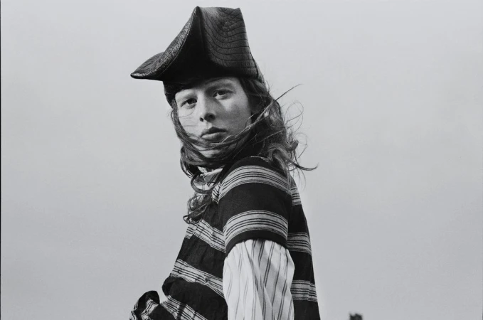 Black and white portrait of a person with long hair wearing a striped shirt and a tricorn hat against a plain, overcast sky.