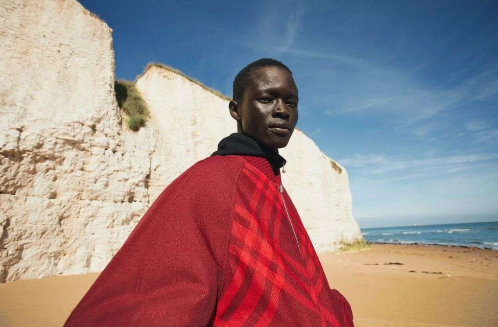 An adult male wearing a maroon patterned jacket stands on a sandy beach with chalk cliffs and blue skies visible in the background.