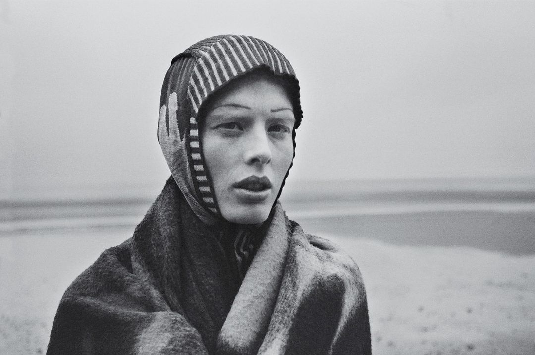 Close-up portrait of a model wearing a striped hood and textured coat outdoors against an overcast sky, by Kerry J Dean.