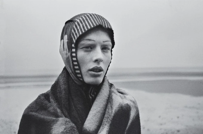 Close-up portrait of a model wearing a striped hood and textured coat outdoors against an overcast sky, by Kerry J Dean.