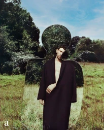Man wearing a black coat standing in a meadow holding an organic-shaped mirror reflecting trees and sky