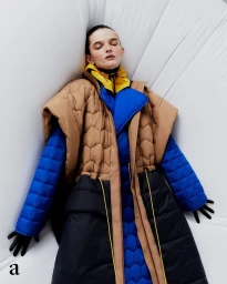 Model in layered puffer coat with camel, blue, and black panels reclining against an inflatable white backdrop
