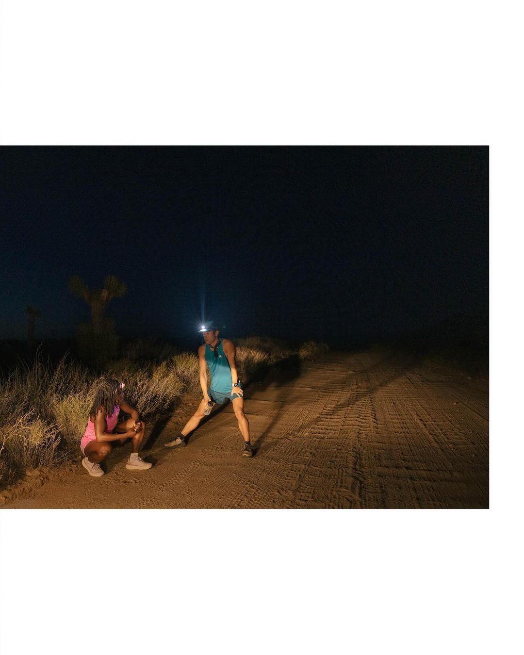 Two women pause on a dirt trail during a night run, lit by headlamps under a dark sky, surrounded by sparse desert vegetation.