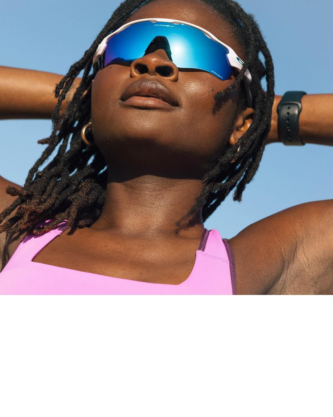 Close-up of an African-American woman with dreadlocks and sunglasses against a blue sky, wearing a pink top and a smartwatch.