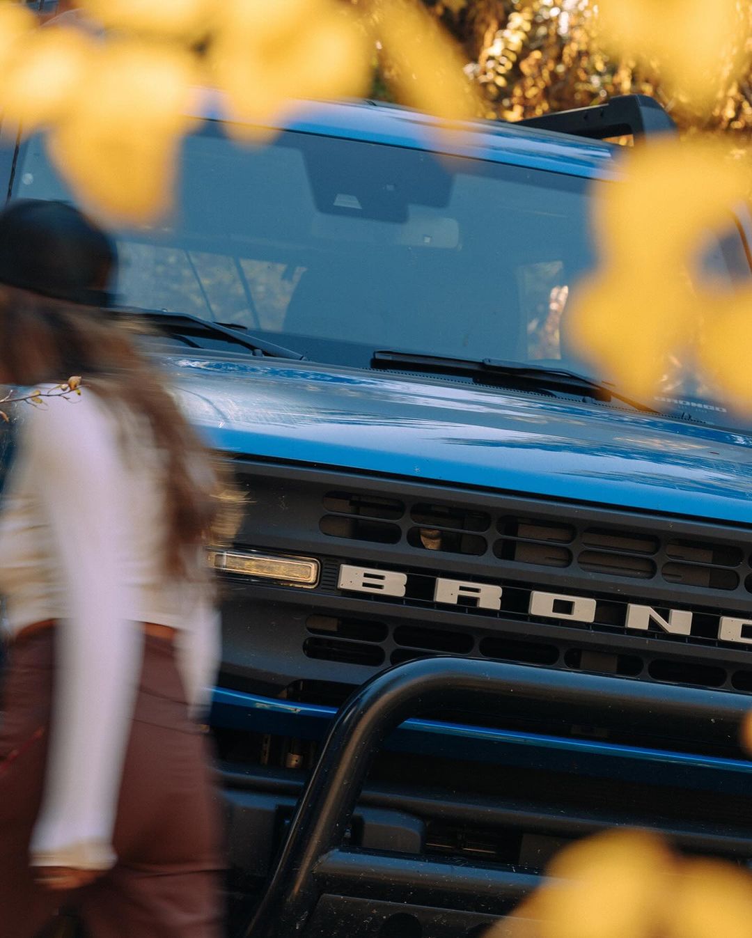 Blue Ford Bronco SUV is partially obscured by out-of-focus yellow leaves. A blurred person walks in the background of a wooded area.