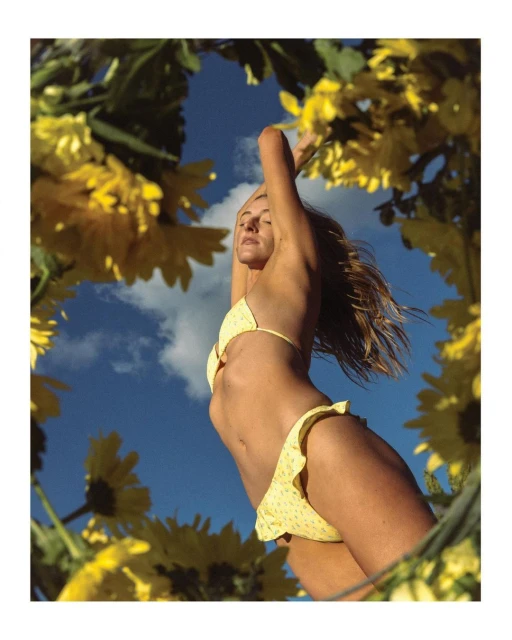 Woman in a yellow bikini posing with arms raised under a bright blue sky framed by yellow daisy-like flowers, shot from a low angle.