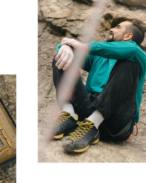 Adult man sits with arms around legs on a rock formation while wearing climbing shoes, black pants and a green sweater.