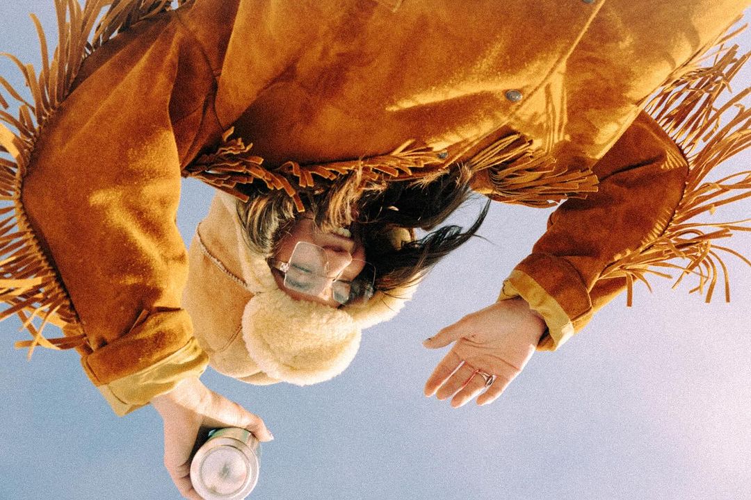 Low angle shot of a person wearing a brown jacket with fringe, glasses, and a trapper hat while holding a metal container.