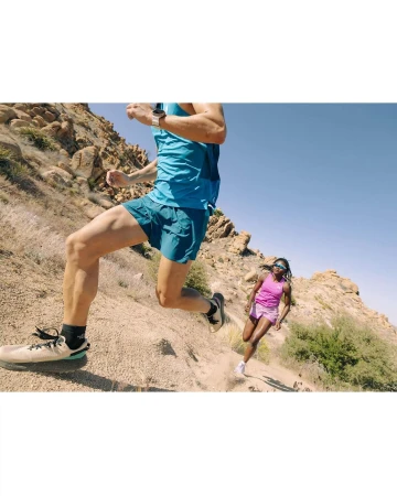 Two athletes trail running downhill in the mountains on a sunny day. The man leads with the woman following, wearing athletic apparel.