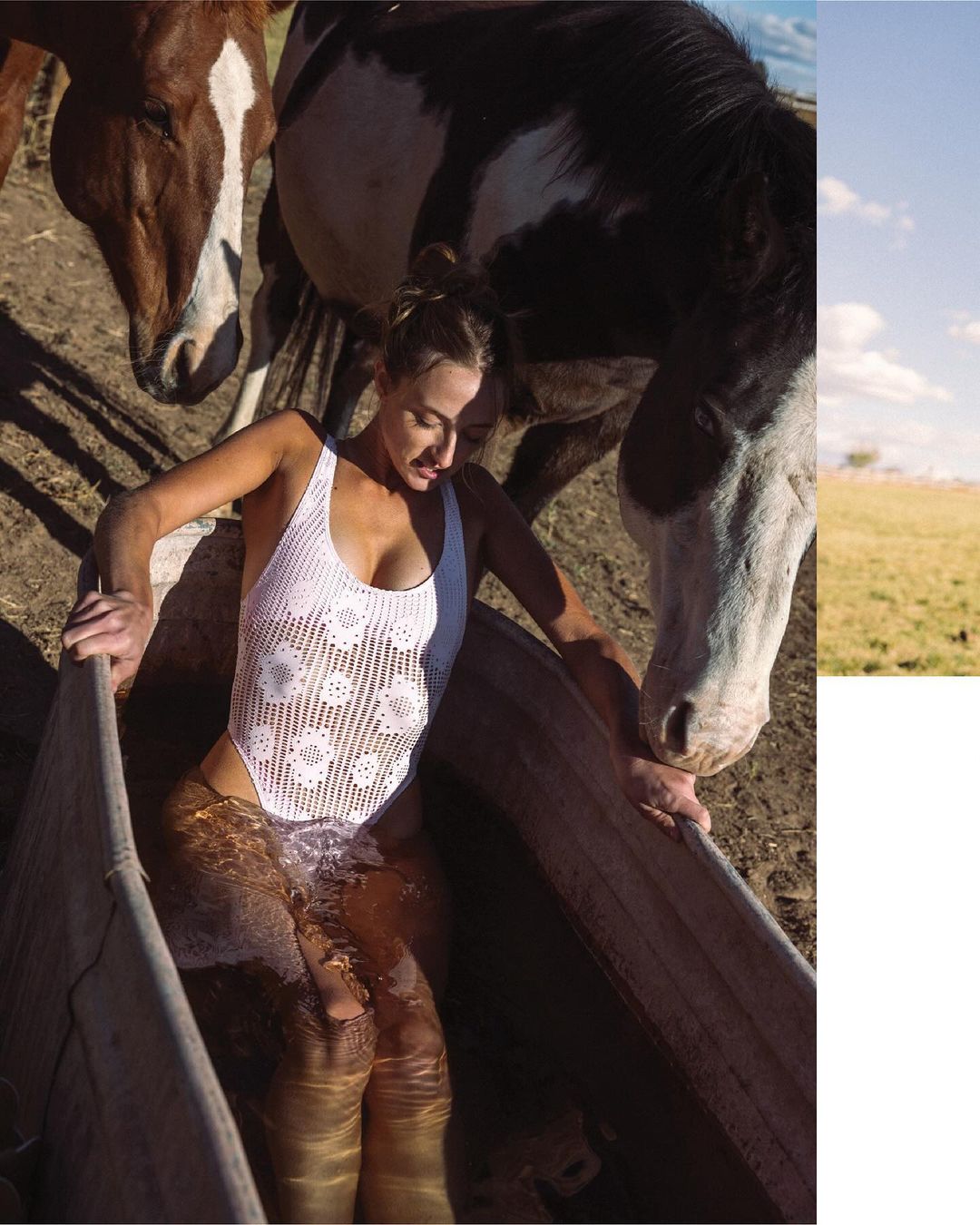 A woman in a white bathing suit is waist-deep in water in a trough surrounded by two horses in an outdoor, rural setting.