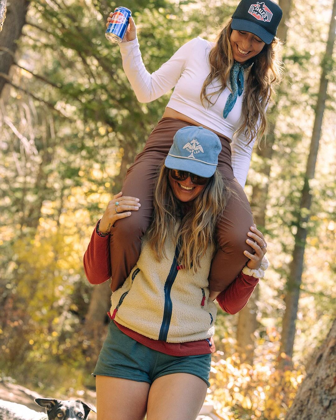 Two smiling women enjoying the outdoors, one giving the other a piggyback ride. One is holding a beverage can. Forest background.