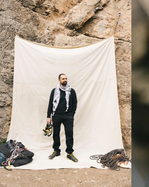 Bearded man in climbing gear poses in front of a canvas with ropes and equipment, set against a rocky mountain backdrop.