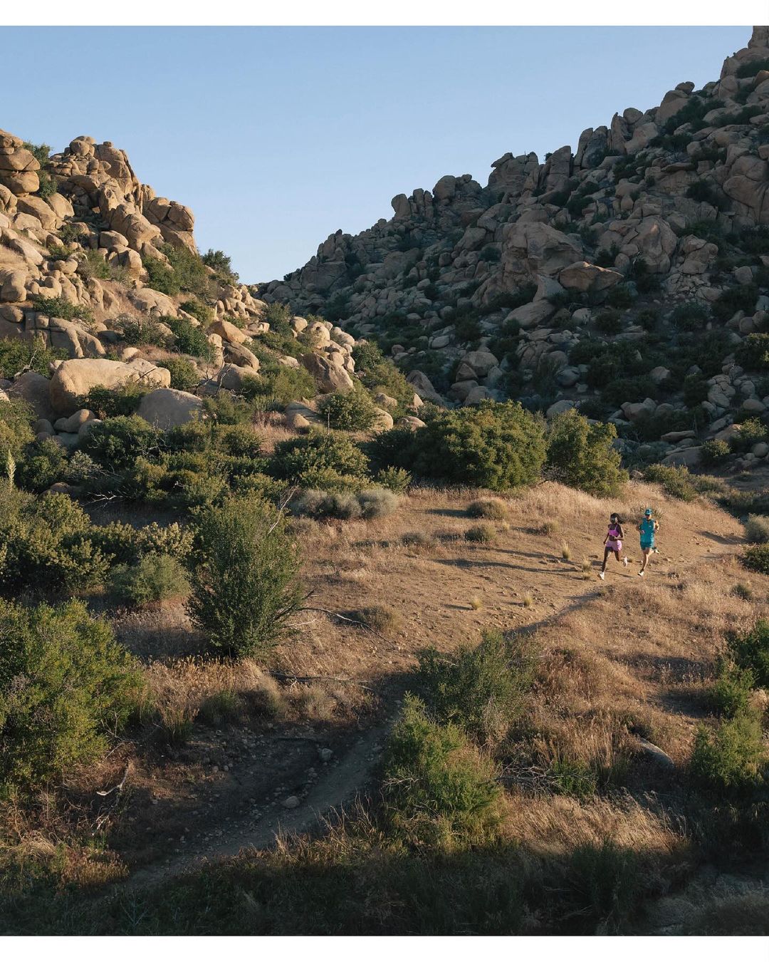 Two adult females trail running on a grassy hillside in a rocky, mountainous landscape under a clear blue sky during daytime.
