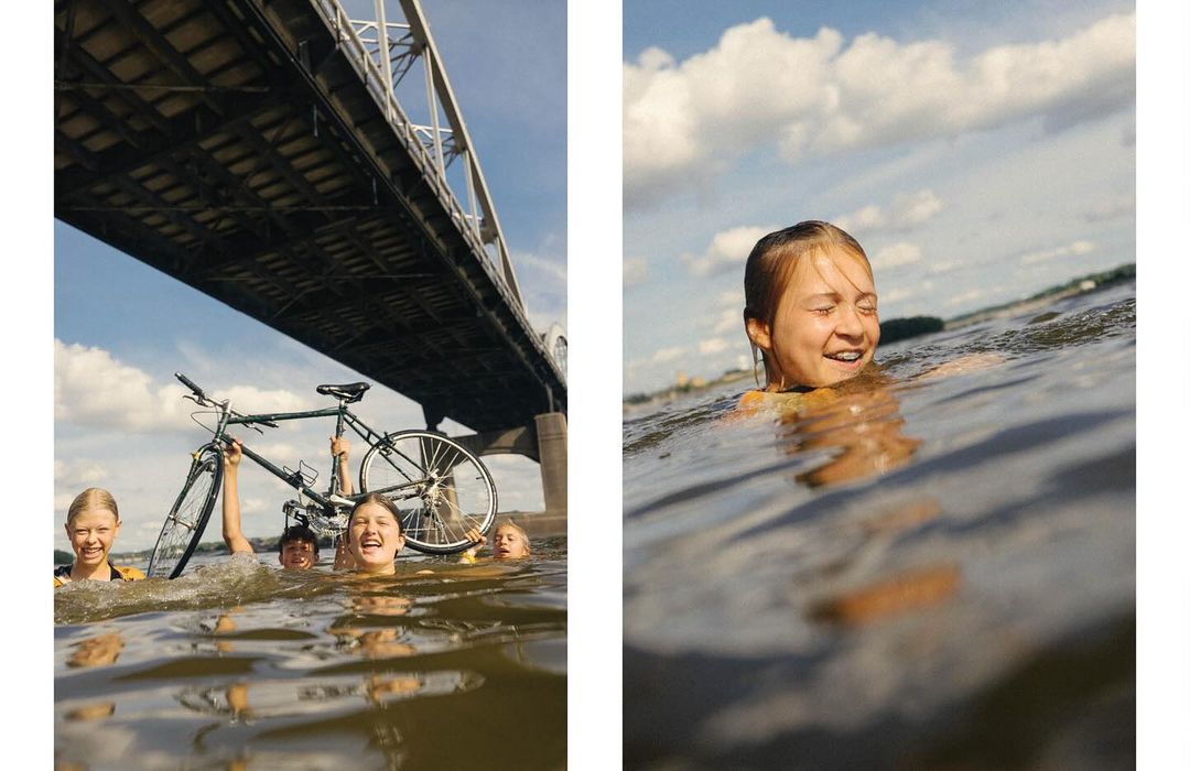 A group of kids with a bicycle are swimming in a river beneath a large bridge on a sunny day with bright, puffy clouds overhead.