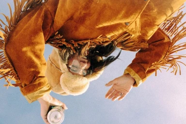 Upside-down portrait of a person wearing a brown fringed suede jacket and a cream trapper hat against a clear blue sky.