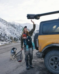 Woman wearing waders holding a fishing net with a baby in a carrier and a dog standing beside an off-road vehicle