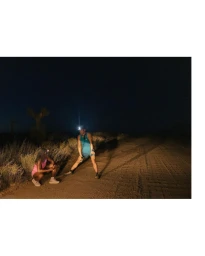 Two runners at night on a dirt path wearing headlamps, with one person stretching and the other crouched nearby in an arid landscape.