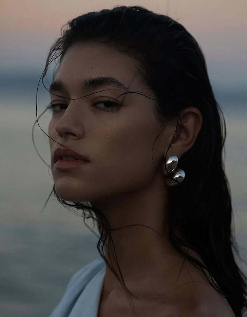 Close-up portrait of a woman with wet dark hair and silver earrings, with a blurred ocean and sky background at dusk.
