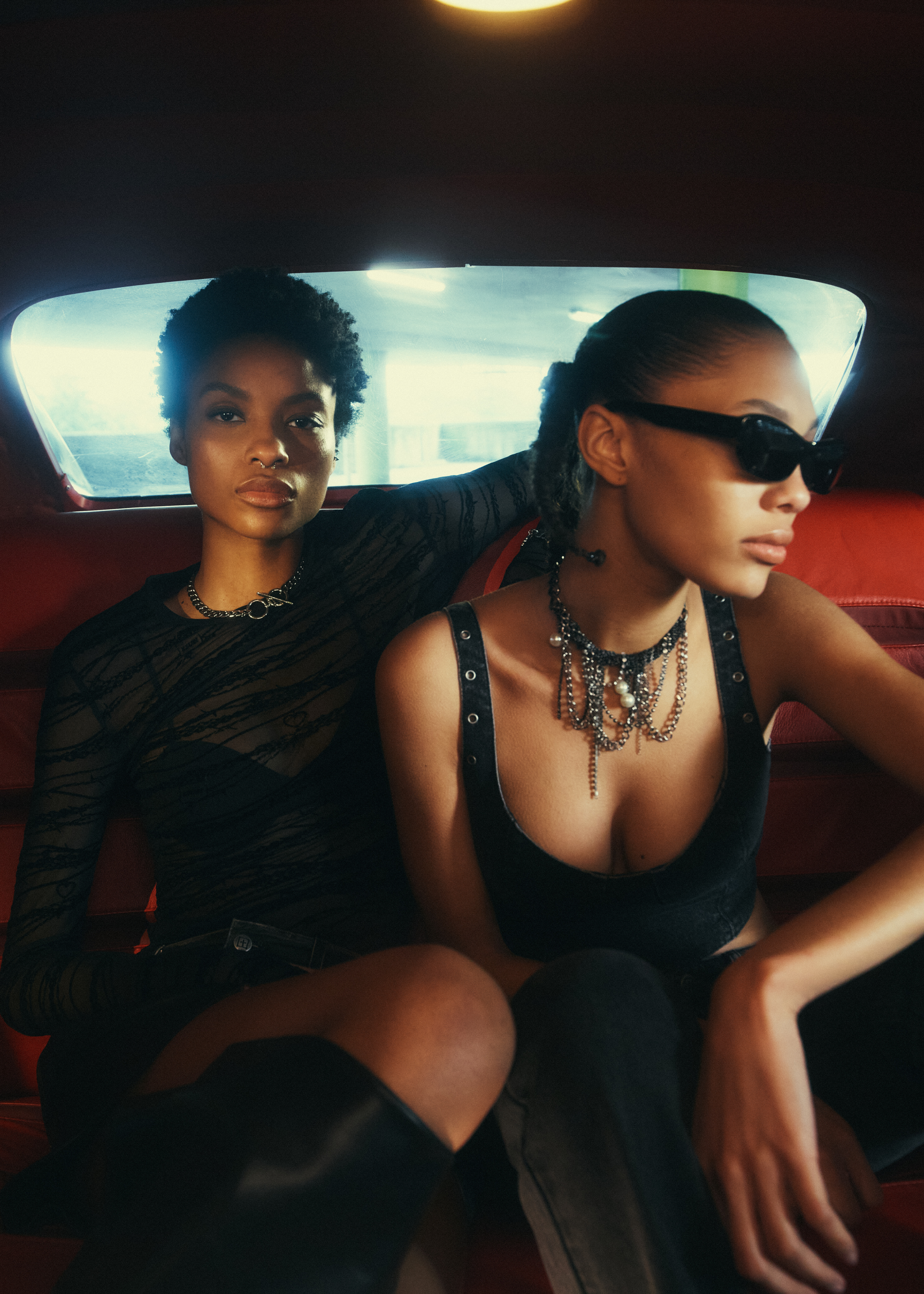 Two stylish adult females pose indoors with sleek black clothing and jewelry, seated closely with a red backdrop and soft lighting.