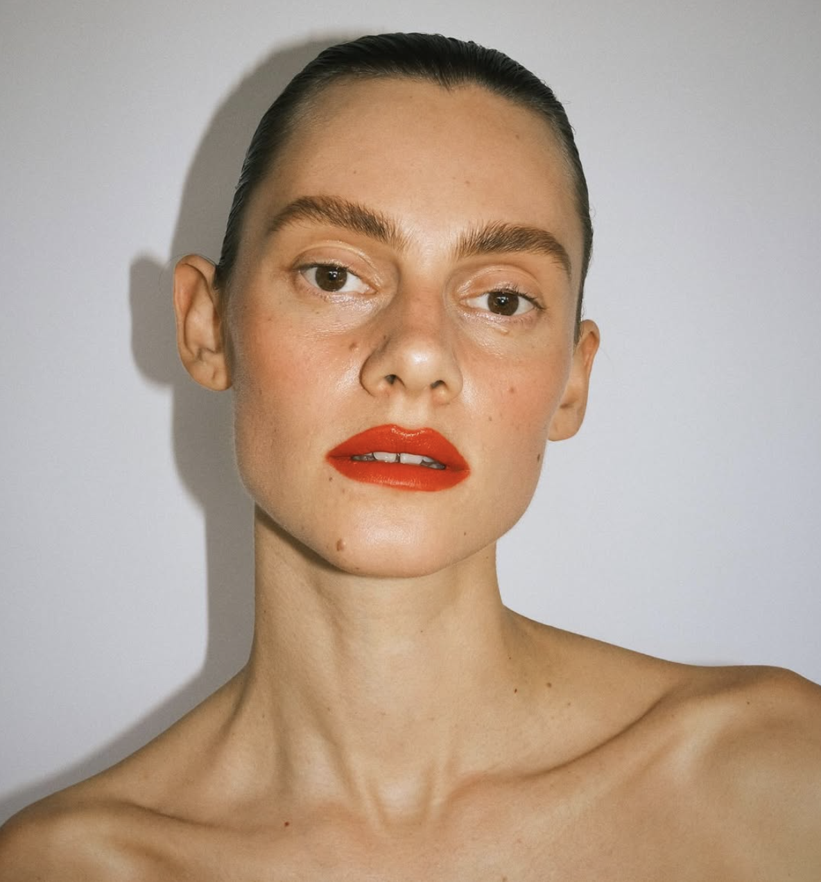 Close-up portrait of a woman with slicked-back hair and bright red lipstick against a bright white background in a photo studio setting.