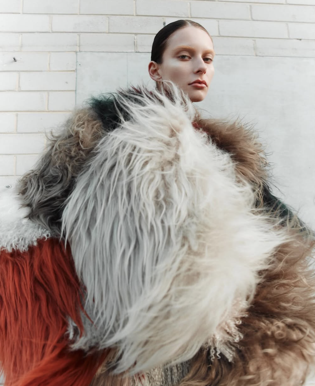 Fashionable model in an oversized multicolor fur coat with a sleek hairstyle posing against a neutral wall in a studio setting.