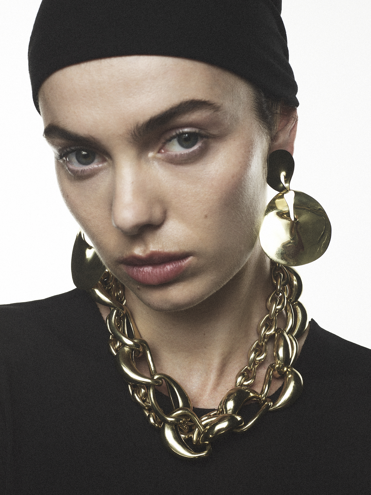 Close-up shot of a woman with a black headscarf wearing gold jewelry against a white background. Beauty shot. Fashion editorial.