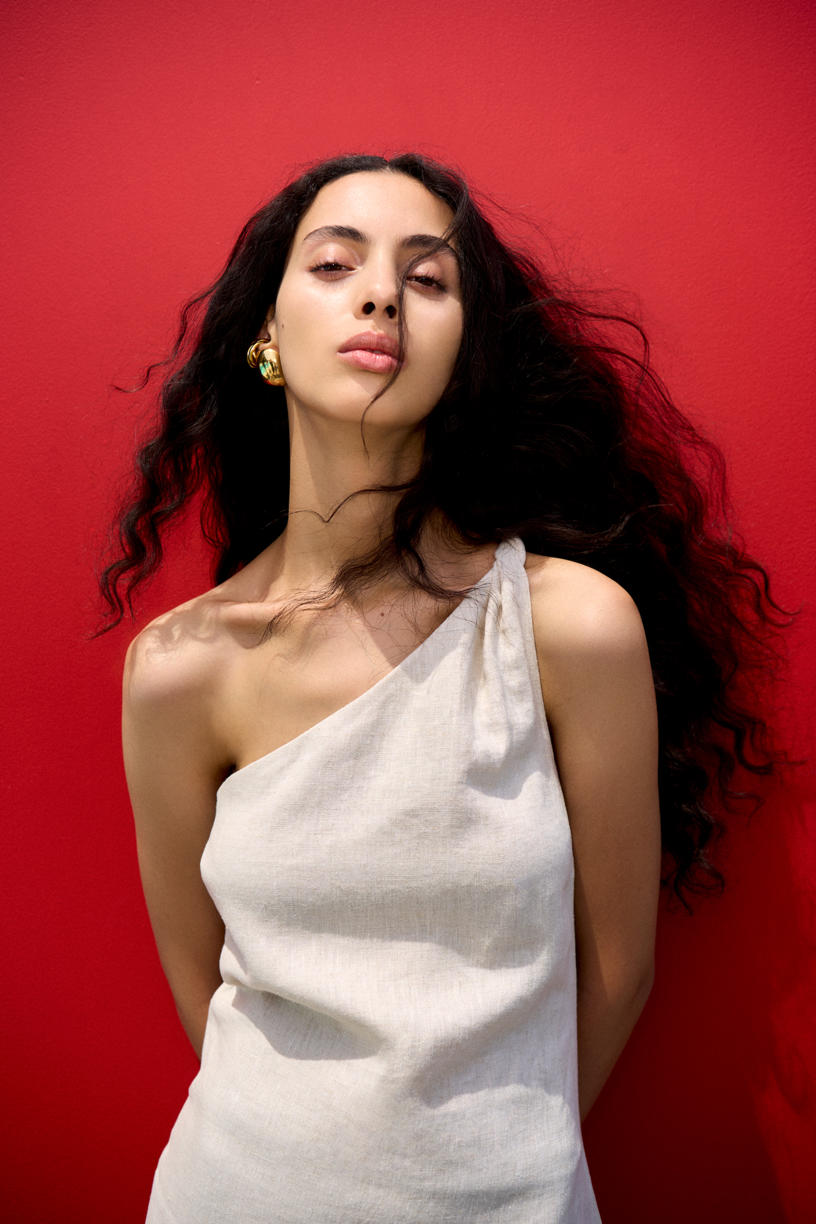 Portrait of a young woman with long, dark, curly hair against a solid red background, wearing a one-shoulder cream-colored linen dress.