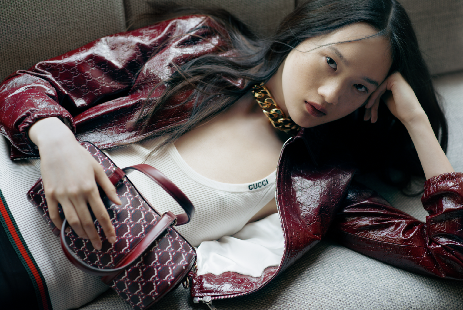 Asian woman in a red jacket, white top, and gold necklace lying on a grey couch with a patterned handbag. Fashion editorial shot.