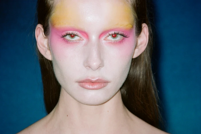 Close-up portrait of a model with vibrant pink and yellow eye makeup and pale skin against a deep blue background.