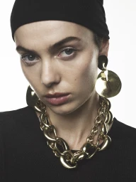 Close-up portrait of a woman wearing a black head covering and large gold statement jewelry including drop earrings and a chunky chain necklace.