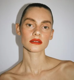 Close-up portrait of a woman with slicked-back dark hair, bare shoulders, and bold red lipstick against a plain white wall