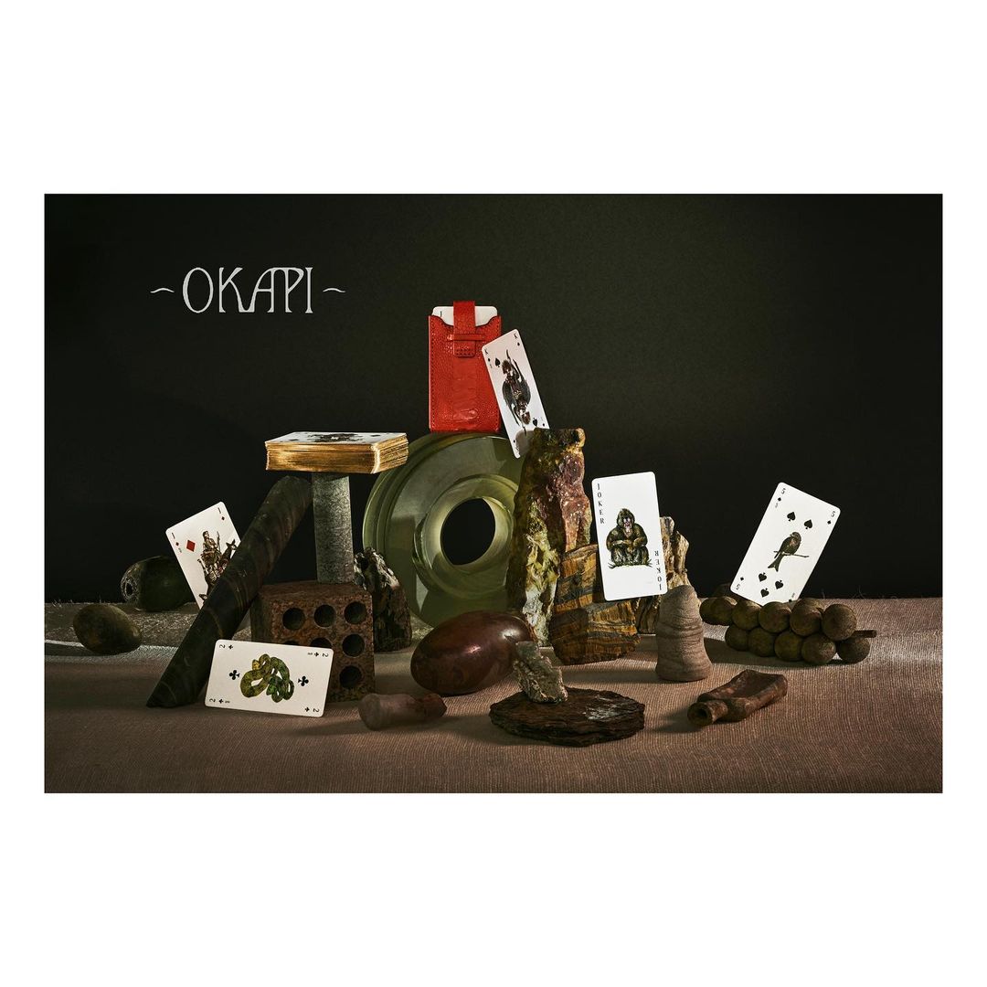 Still life composition of playing cards, packaging, and sculptural objects arranged on a textured surface against a dark backdrop.