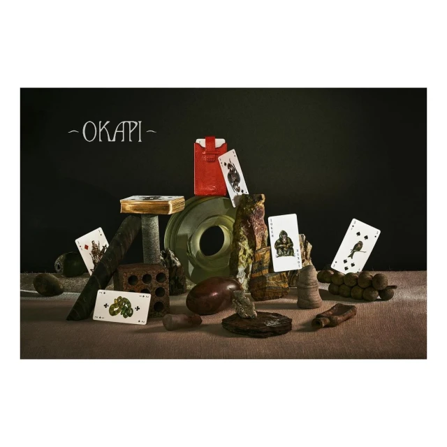 Still life composition of playing cards, packaging, and sculptural objects arranged on a textured surface against a dark backdrop.