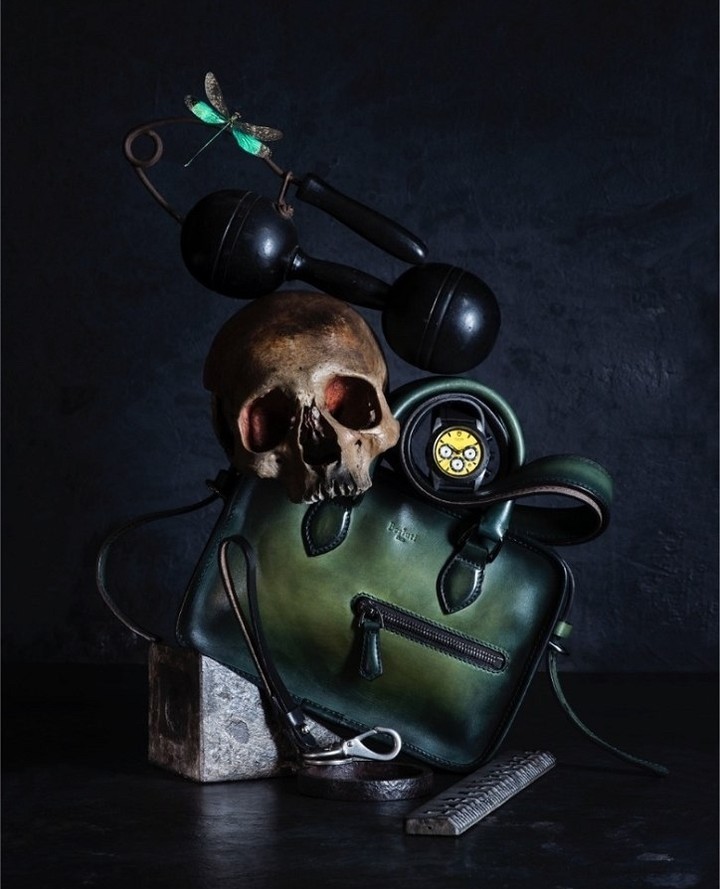 Studio shot featuring a green leather bag, skull, dumbbells, watch winder, and dragonfly against a dark backdrop, creating a contrast.