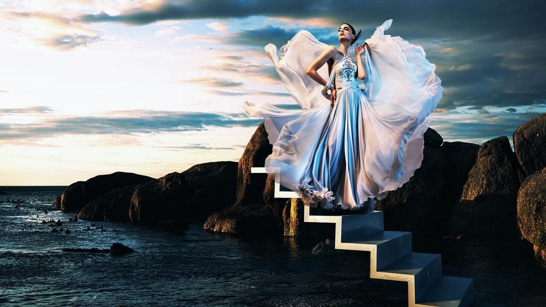 A woman in a flowing gown stands on stairs leading from rocks to the ocean under a dramatic sky with clouds at sunset.