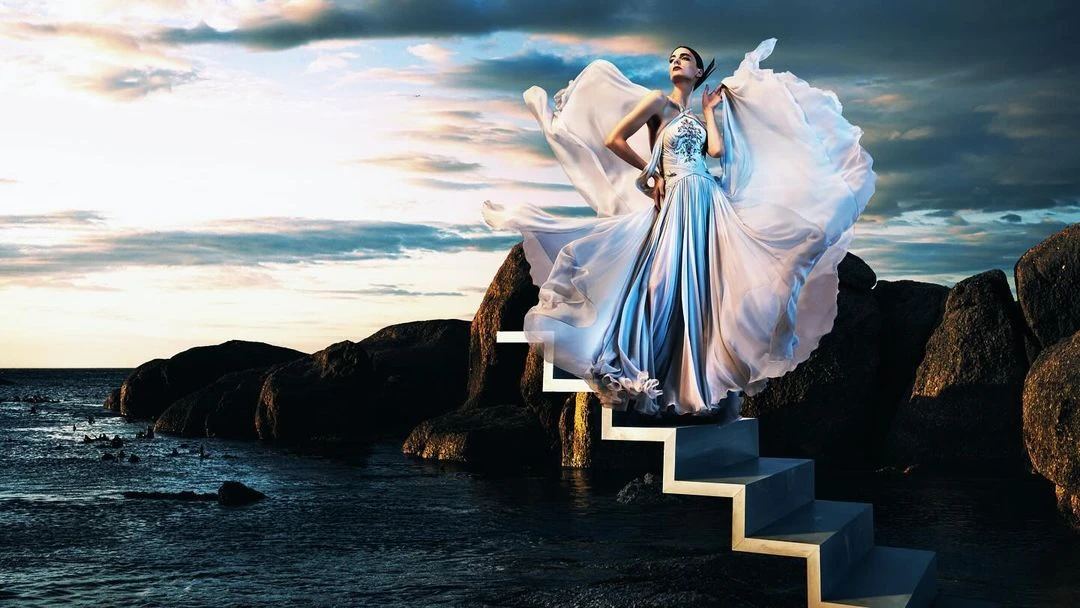 A woman in a flowing gown stands on stairs leading from rocks to the ocean under a dramatic sky with clouds at sunset.