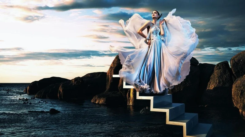 A woman in a flowing gown stands on stairs leading from rocks to the ocean under a dramatic sky with clouds at sunset.