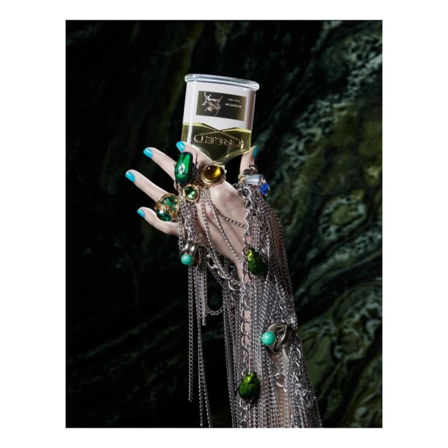 Hand adorned with jewelry and turquoise nail polish holds Creed perfume against a dark green marble backdrop in a beauty shot.