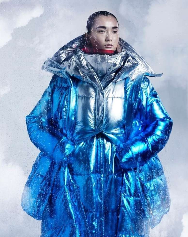 Asian adult model wearing a metallic blue oversized puffer jacket with an exaggerated collar against a blurred cloudy background.